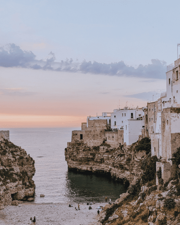 Coastal cliffs and historic buildings at sunset, with a serene beach below, in Polignano a Mare, Italy.